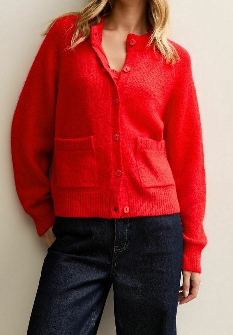Femme portant un cardigan rouge vif boutonné avec des poches frontales et un jean bleu foncé, debout devant un fond neutre.