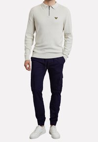 Polo en maille beige clair avec col zippé et petit logo, associé à un pantalon cargo bleu marine et des baskets blanches. Comprend des manches longues et une ceinture ajustable.