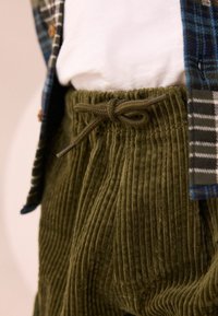 Olivgrüne Cordhosen mit vertikalen Rippungen, ausgestattet mit einem Kordelzugverschluss und getragen mit einem karierten Hemd und einem weißen Shirt.