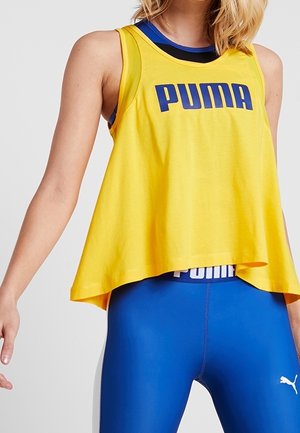 Mujer con una camiseta sin mangas amarilla holgada de Puma sobre un top deportivo azul y leggings azules de Puma con el logo blanco en el muslo izquierdo.