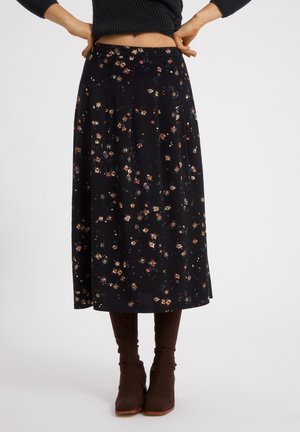 Kvinde iført en sort midi-nederdel med små blomstermønstre, kombineret med brune hælede støvler, står mod en ensartet hvid baggrund.