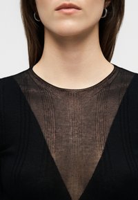 Primer plano de una persona con una blusa negra que tiene un panel en forma de V translúcido y estampado en el pecho, y pendientes de aro plateados.