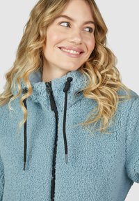 Veste en polaire bleu clair avec une surface texturée, fermeture éclair sur le devant et cordons noirs autour du cou. Design doux et décontracté.