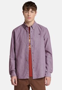 Camisa de botón de color púrpura claro con cuello y mangas largas, con un pequeño logotipo en el pecho. Lleva puesta sobre una camiseta roja.