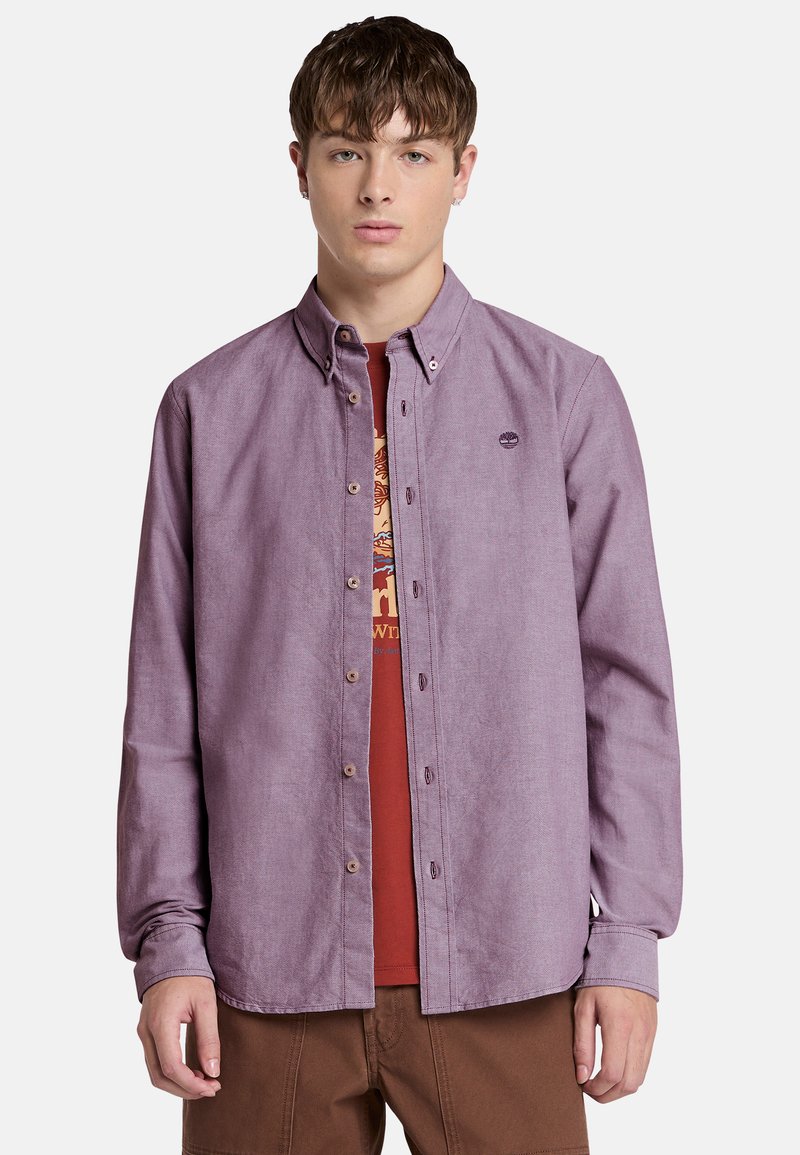 Camisa de botón de color púrpura claro con cuello y mangas largas, con un pequeño logotipo en el pecho. Lleva puesta sobre una camiseta roja.