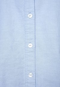 Ljusblå skjorta med knapp fram, tillverkad i tyg med strukturerad väv, med silverfärgade knappar och kontrasterande sömmar längs knapplistan.