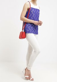 Top in pizzo blu reale con spalline bianche, abbinato a pantaloni skinny bianchi e sandali con tacco bianco. Una borsa rossa aggiunge un dettaglio di contrasto.