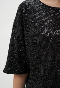 Top nero in paillettes con vestibilità ampia, scollatura rotonda e maniche corte. Presenta una superficie testurizzata e scintillante con elementi riflettenti.
