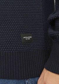 Navy blue knitted sweater with textured pattern, ribbed cuffs and hem. Black fabric label at bottom with "JACK & JONES" and established date.