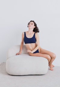 Tummansininen croptop ja bikinit yhdessä vaaleanbeigeillä, pehmeillä, kaarevilla sohvilla minimalistisessa ympäristössä. Sujuva pinta, yksinkertainen muotoilu.
