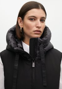 Musta puffer-liivi korkealla kauluksella, joissa on kiiltävä kangas, vetoketjukiinnitys ja logoyksityiskohdat hihnoissa. Sileä tekstuuri, moderni muotoilu.