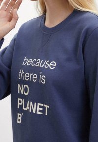 Persona con sudadera azul que lleva el texto blanco "porque NO HAY UN PLANETA B" en el frente, levantando su mano izquierda cerca de su cara.
