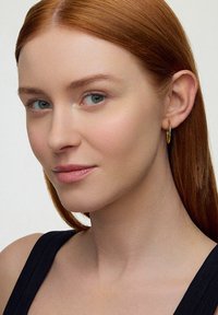 Złota kolczyka w kształcie obręczy noszona na prawym uchu, o gładkim, błyszczącym wykończeniu. Modelka ma długie czerwone włosy, ubrana w czarną bluzkę o prostym wzorze.