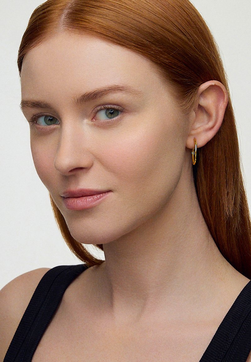 Złota kolczyka w kształcie obręczy noszona na prawym uchu, o gładkim, błyszczącym wykończeniu. Modelka ma długie czerwone włosy, ubrana w czarną bluzkę o prostym wzorze.