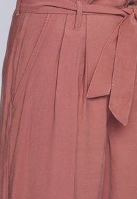 Pantalons larges en coton rose avec plis sur le devant, une ceinture en tissu et des poches latérales. Texture lisse avec des détails de couture visibles.