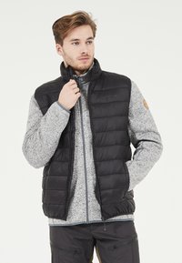 Svart pufferväst med hög krage över en gråmelerad zip-up fleecejacka. Västen har ett kviltat mönster och sidofickor.
