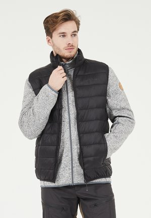 Sort puffervest med høj krave over en grå spraglet lynlås-fleecejakke. Vestens design er quiltet og har sidelommer.