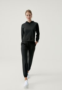 Felpa nera con cappuccio, coulisse e tasca frontale, abbinata a pantaloni jogger neri a sigaretta. Il tessuto sembra liscio e leggero. Scarpe bianche senza lacci.