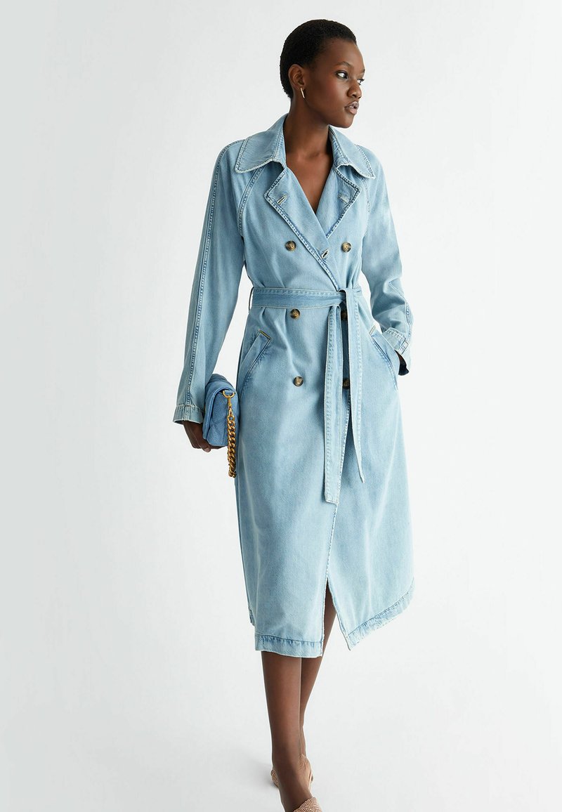 Trench coat in denim azzurro con design doppiopetto, grande colletto, cintura a fiocco e spacchi frontali; presenta bottoni dorati e tasche.