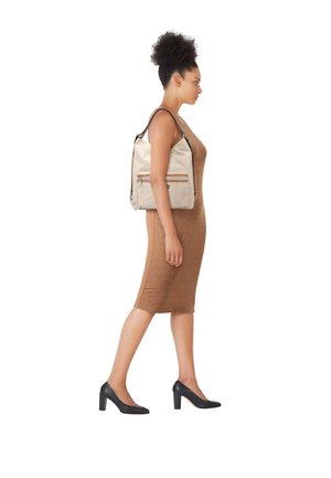 Mujer caminando con un vestido marrón sin mangas, tacones negros y llevando un bolso de hombro beige con cremalleras delanteras.