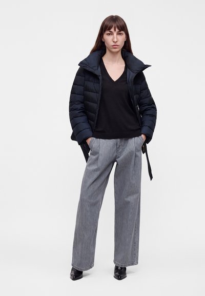 Femme portant une veste matelassée noire par-dessus un pull noir à col en V, un pantalon gris ample et des chaussures noires pointues, debout les mains dans les poches.