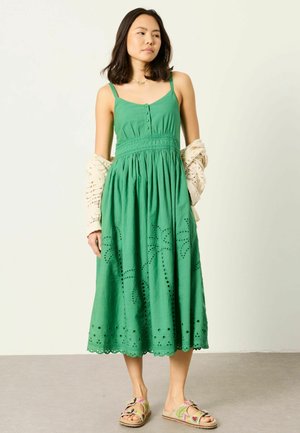 Kvinde i grøn ærmeløs midikjole med hulmønsterdetaljer, der holder en beige strikket cardigan, iført blomstrede sandaler, stående på gråt gulv.