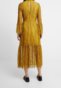 Vestido amarillo de encaje con mangas largas acampanadas, abertura trasera en forma de ojal y dobladillo en capas. Presenta patrones florales intrincados y un acabado texturizado.