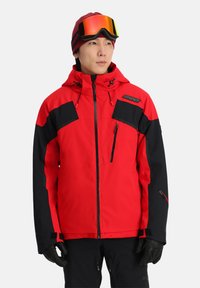 Rote und schwarze Skijacke mit einer Reißverschlusstasche auf der Brust, verstellbarer Kapuze, elastischen Bündchen und einem Logodetail. Kombination mit schwarzen Handschuhen.