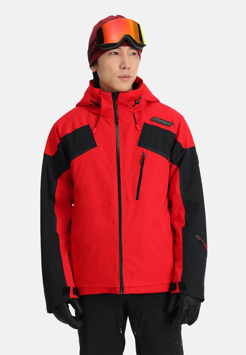 Rote und schwarze Skijacke mit einer Reißverschlusstasche auf der Brust, verstellbarer Kapuze, elastischen Bündchen und einem Logodetail. Kombination mit schwarzen Handschuhen.