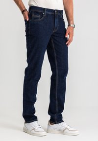 Jeansy z ciemnoniebieskiego denimu o prostym kroju, z pomarańczowym przeszyciem, przednimi kieszeniami i zapięciem na zamek błyskawiczny. Noszone z białymi tenisówkami.