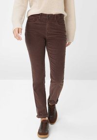 Pantaloni in velluto a coste marroni con un design aderente, dotati di tasche anteriori e chiusura con bottone, abbinati a sneakers marroni scure.
