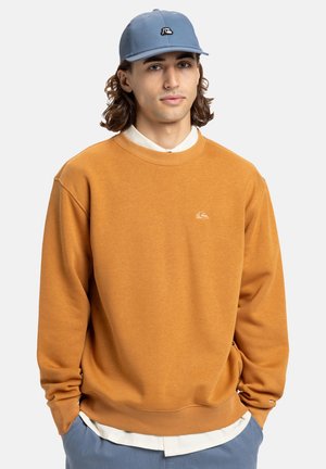 Oranje sweatshirt met een ronde hals en ribgebreide boorden, gecombineerd met een lichtblauw pet met een logo. Model staat tegen een effen achtergrond.