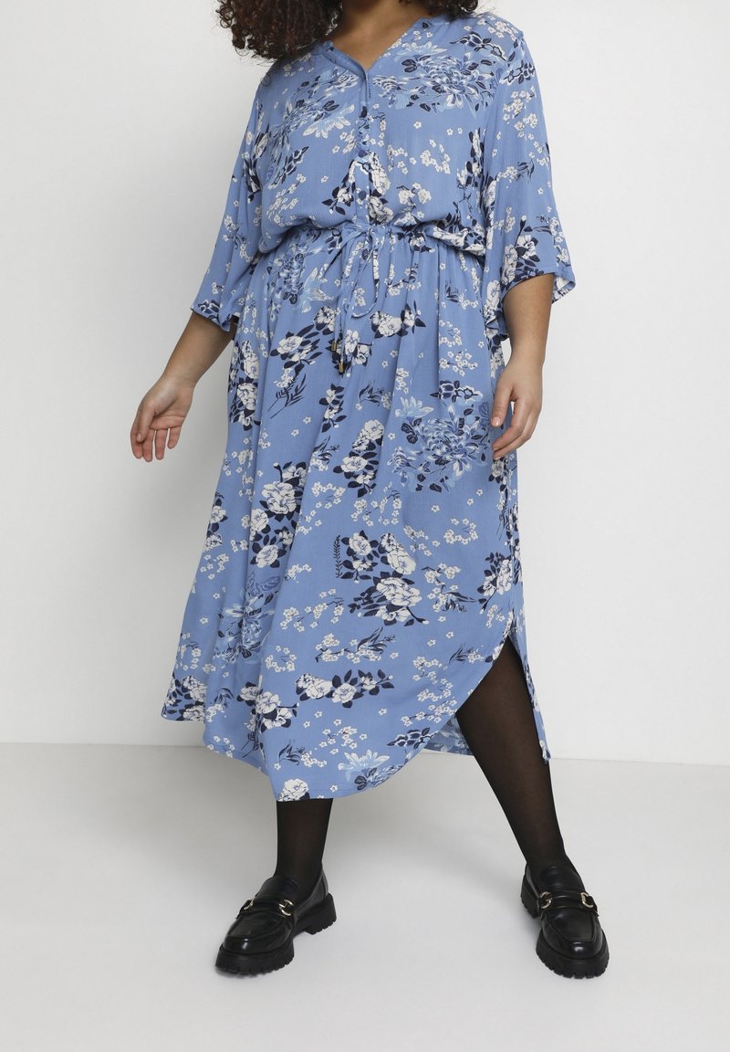 Femme portant une robe midi bleue à motifs floraux avec taille à cordon, des collants noirs et des mocassins noirs, debout devant un fond blanc.