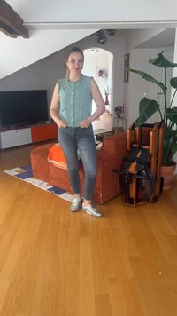 Νέα γυναίκα με καθημερινά ρούχα στέκεται σε ένα σαλόνι με πορτοκαλί καναπέ, μεγάλο φυτό, τηλεόραση και ξύλινη μηχανή κωπηλασίας.
