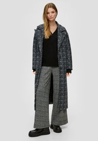 Langer, gestreifter Mantel in Schwarz und Weiß mit breiten Revers, kombiniert mit einem schwarzen Strickpullover und grauen, strukturierten weiten Hosen. Schwarze Plateauschuhe.