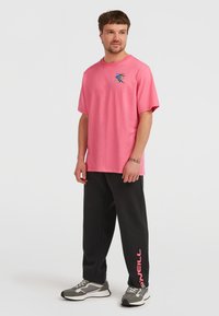 Lyserød bomulds t-shirt med en grafisk design på venstre bryst, parret med sorte sweatpants med et lyserødt logo på siden. Grå sportsko.