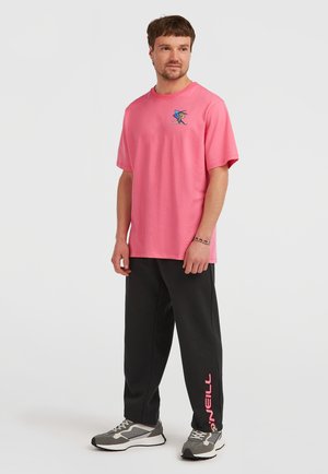 T-shirt en coton rose avec un imprimé graphique sur la poitrine gauche, associée à un pantalon de survêtement noir portant un logo rose sur la jambe.