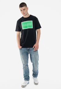 Black cotton t-shirt featuring a large green “oasis” graphic print. Worn with light blue jeans and white sneakers. Simple, casual design.