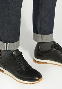 Black athletic shoes made of textured leather with a smooth finish, featuring a white sole and tan accents, paired with dark denim pants.