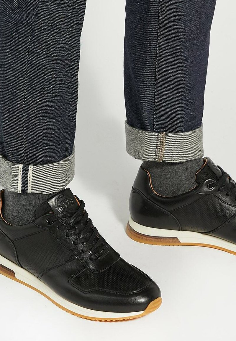 Black athletic shoes made of textured leather with a smooth finish, featuring a white sole and tan accents, paired with dark denim pants.