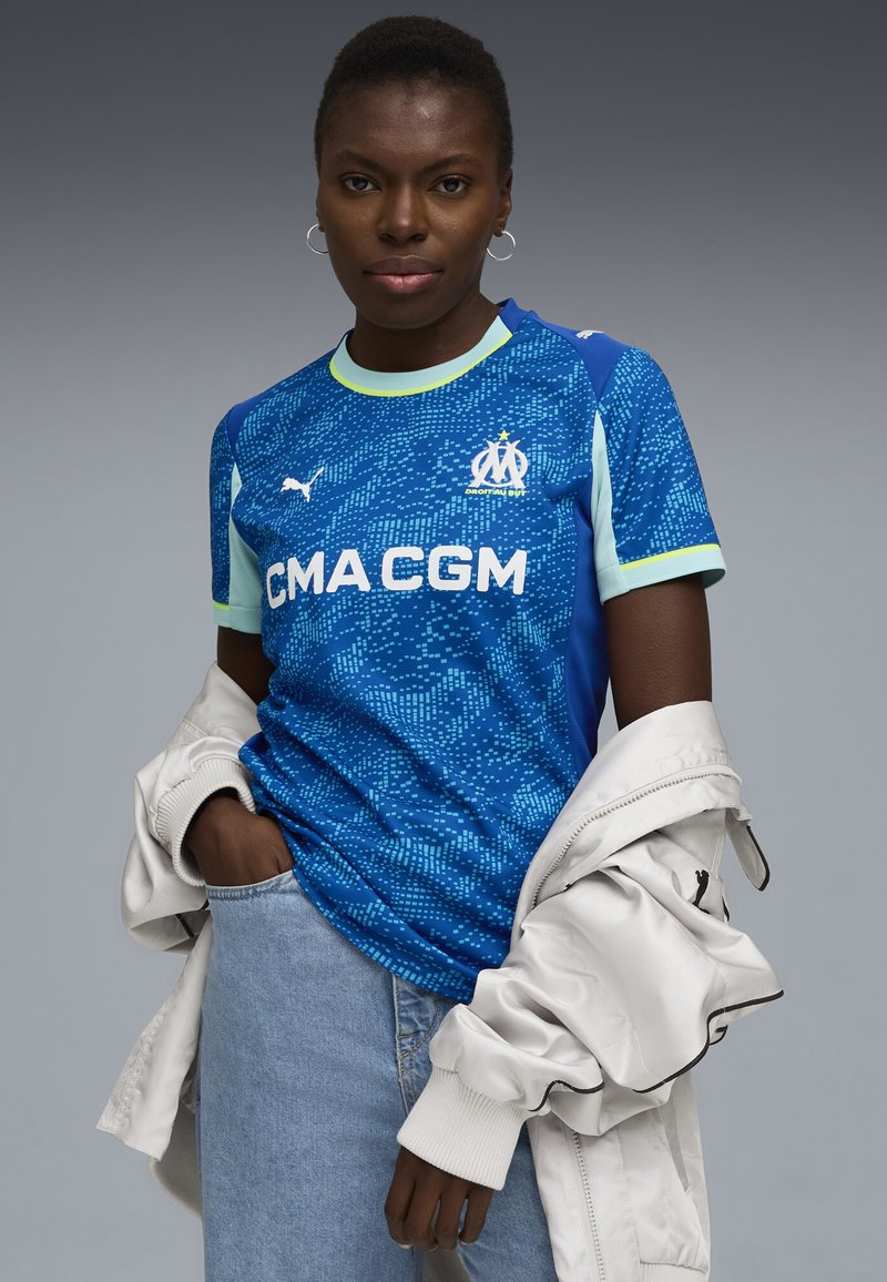 Młoda kobieta w niebieskiej koszulce piłkarskiej Marsylii, w jasnoniebieskich dżinsach i białej kurtce zarzuconej na ramiona, pozująca na szarym tle.