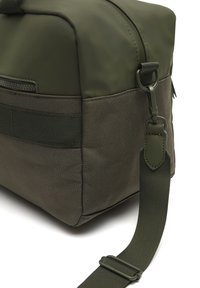 Zelená nylonová taška s lesklým povrchom, s textúrovaným dnom, zipsom a nastaviteľným popruhom. Obsahuje detail bočného webbing.