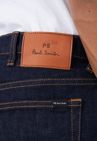 Detailní záběr tmavě modrých džínů z denimu s oranžovým šitím a hnědou koženou štítkem s nápisem "PS Paul Smith" na pásku.