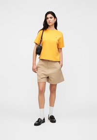 Κίτρινη βαμβακερή μπλούζα, μπεζ σορτς, μαύρο τσαντάκι ώμου, λευκές κάλτσες και μαύρα παπούτσια. Απλός σχεδιασμός χωρίς ορατά μοτίβα ή διακοσμητικά στοιχεία.