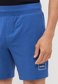 Plave pamučne kratke hlače s elastičnom trakom u struku, bočnom džepom i bijelim pravokutnim logotipom na lijevoj nozi. Mekana tekstura i ležeran dizajn.