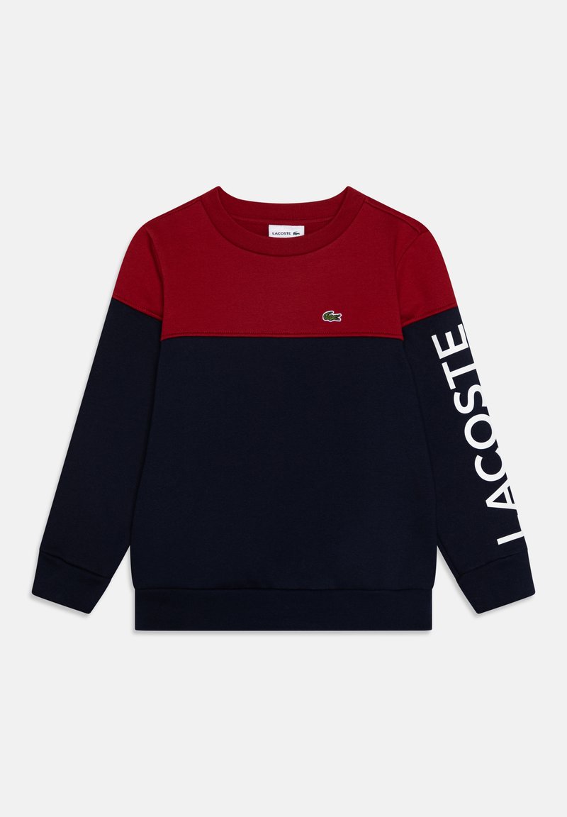 Sweatshirt avec une section supérieure rouge et une section inférieure bleu foncé, avec un logo "LACOSTE" blanc sur la manche et un petit emblème de crocodile vert.