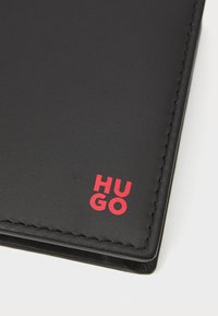 Schwarzes Lederportemonnaie mit rotem "HUGO"-Logo. Merkmale sind klare Linien, sichtbare Nähte und eine glatte Textur. Kompaktes, rechteckiges Design.