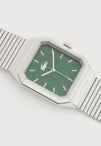 Ασημένιο ορθογώνιο ρολόι χειρός με πράσινο καντράν, ασημένιους δείκτες ώρας και δείκτες, κόκκινη άκρη στον δείκτη των δευτερολέπτων και μεταλλικό μπρασελέ, εμφανές το λογότυπο Lacoste.