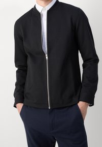 Homme portant une veste noire à fermeture éclair par-dessus une chemise blanche à boutons avec un pantalon bleu marine, mains dans les poches, devant un fond uni.