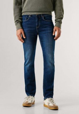 Uomo con jeans slim fit blu scuro, maglione verde oliva e sneaker bianche in piedi su sfondo uniforme.